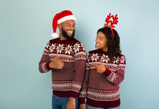New Year 2022 celebration. Young black couple in Christmas sweaters pointing index fingers at each other and smiling, posing on blue background, free space