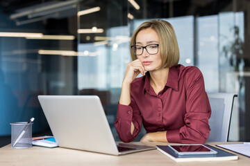 Professional businesswoman in glasses thinking deeply while working on her laptop, analyzing data...