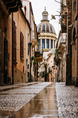 Back Side Of Duomo Di San Giorgio In Ragusa, Sicily: Narrow Historic Street Leading Toward Baroque Cathedral Dome And Stone Facades