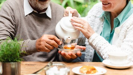 Senior couple enjoy afternoon tea outdoors, pouring tea. Senior couple man and woman relax in garden with tea and snack. Relaxed senior couple drink tea with snack in garden, afternoon relax