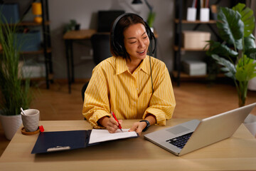 Asian woman using headset and laptop for a video call while writing notes in her home office