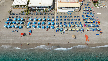 Aerial view of an organized beach club featuring neat rows of light blue parasols. The composition captures the transition from the italian beach resort to the gentle white surf of the turquoise sea.