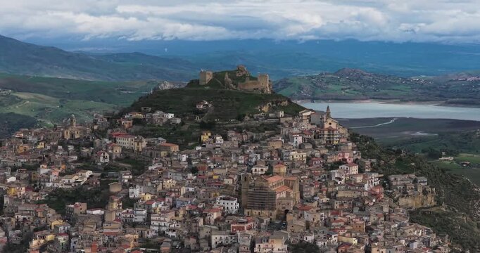 Aerial view of Agira's ancient architecture clustered on a hill crowned by a weathered castle, contrasting the earthy tones with the distant blue mountains, Agira, Sicilia, Italy.