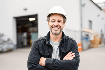 A smiling construction worker in a hard hat stands confidently with arms crossed in an industrial setting.