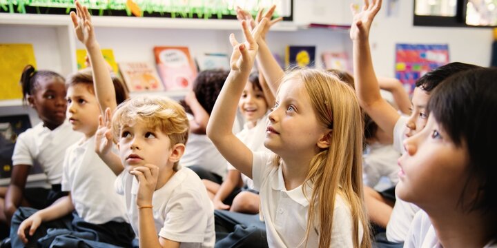 Diverse group of young students, boys and girls, sit on the floor in a classroom raising hands. Diverse students raise their hands, engaged in a classroom activity. Students raise hand in classroom.