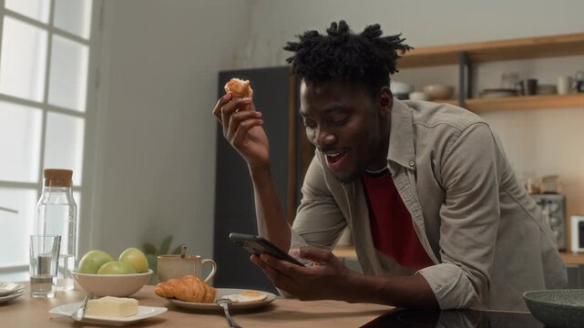 Medium shot of young Black man leaning on kitchen counter and looking at smartphone screen while having croissant and eggs for breakfast, scrolling newsfeed or ordering delivery