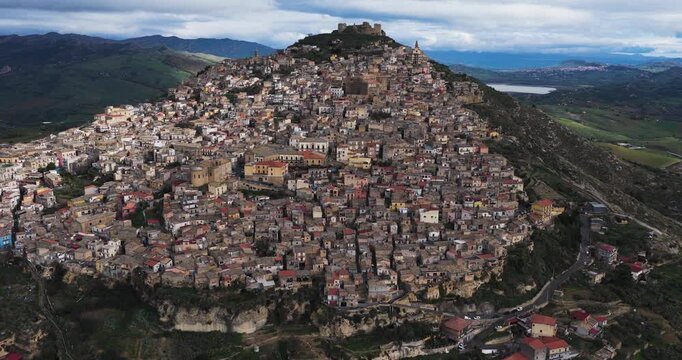 Aerial view of the ancient town of Agira cascading down a steep hillside, crowned by castle ruins, with a backdrop of rolling green hills, Agira, Sicilia, Italy.