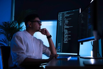 Focused software developer working late at night in dark office. Professional programmer coding on vertical monitor with blue neon light. Cybersecurity expert analyzing data alone.
