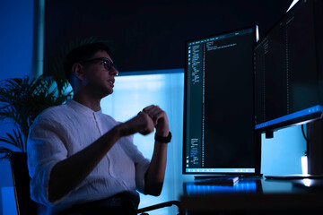 Focused software developer working late at night in dark office. Professional programmer coding on vertical monitor with blue neon light. Cybersecurity expert analyzing data alone.