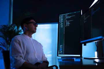 Focused software developer working late at night in dark office. Professional programmer coding on vertical monitor with blue neon light. Cybersecurity expert analyzing data alone.