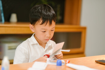 Seven-year-old Asian boy sitting at a desk and focusing on paper cutting for handicrafts in a cozy home setting