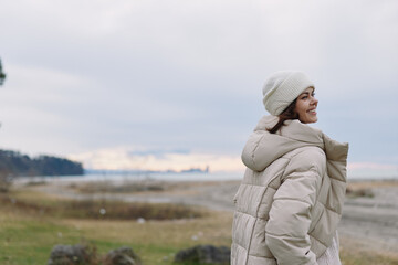 Woman in a beige puffer coat and beanie gazes toward a windy seaside landscape with a calm horizon, creating a serene outdoor fashion portrait.