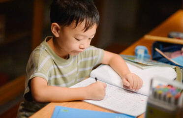 Seven-year-old Asian boy focused on doing homework and writing at a desk in a cozy home setting