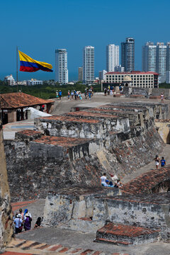 View of weathered stone walls of a historic fort contrast with modern skyscrapers under a clear blue sky, all under the watchful eye of the Colombian flag, Cartagena, Bolivar, Colombia.