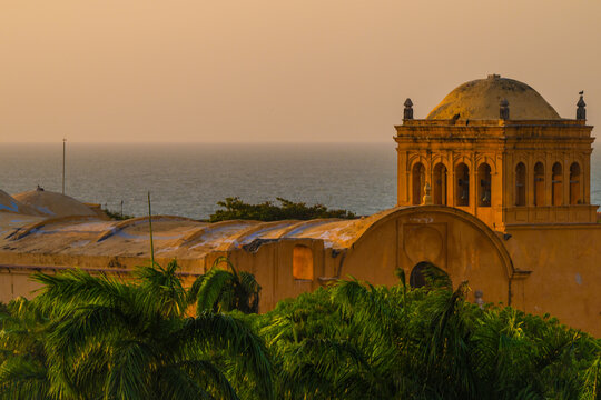 View of golden light bathes the ancient architecture, as seen from ground level, with palm trees against the backdrop of the sea, Cartagena, Bolivar, Colombia.