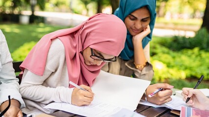 Two women in hijabs study outdoors, focused on writing. Hijabs, study, and writing are central. Women, hijabs, and study in a park setting. Diverse students doing homework together.