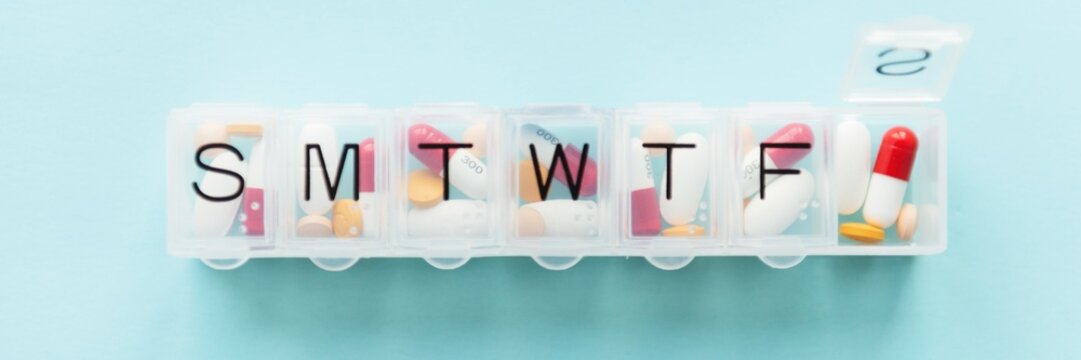 Weekly vitamin pill organizer with compartments labeled S, M, T, W, T, F, filled with various pills. Medicine, pills, and organizer on a light blue background. Vitamins in a box.