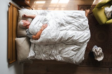 Elderly man sleeping in bed, covered with a white blanket. Peaceful rest, cozy bedroom setting. Overhead view of a sleeping man in a serene environment. Elderly man sleeping in bed.