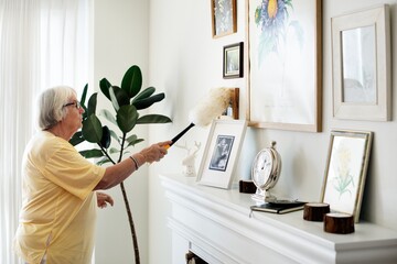 Senior woman dusting picture frames on a mantel. Indoor cleaning scene with a feather duster....