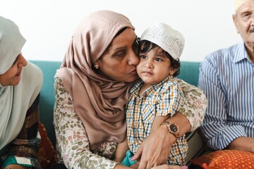 Elderly woman in a hijab lovingly holds a young child wearing a cap. Family gathering with diverse...