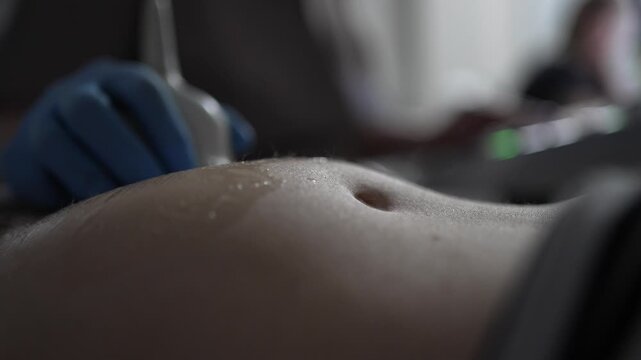 Close-up of a doctor performing an ultrasound examination on a pregnant woman's belly, moving the transducer with gel on her abdomen to monitor the baby's health during a prenatal checkup