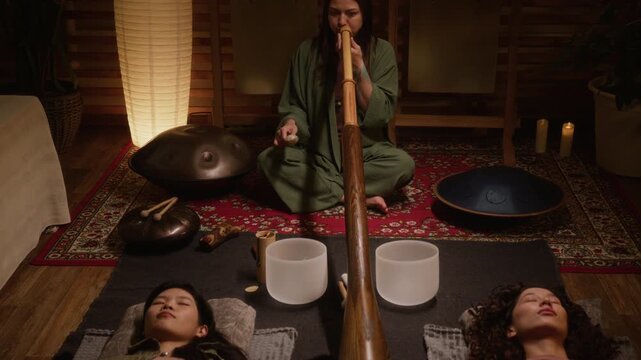 Medium full shot of young Caucasian female sound healing master playing didgeridoo and shaking rattle while two female guests are lying on carpet with closed eyes and meditating