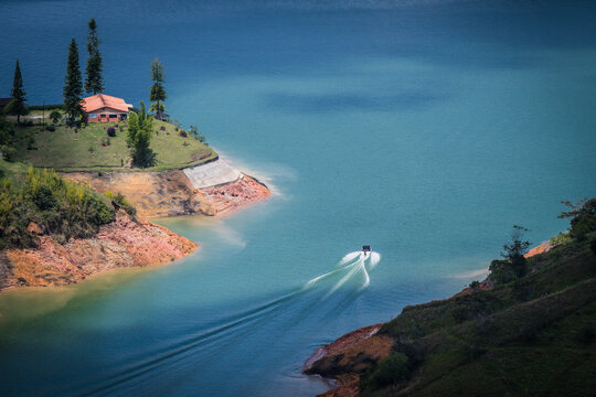 View of a speedboat slicing through the turquoise waters near a red-roofed house on a verdant peninsula, creating a dynamic contrast of colors, Guatape, Antioquia, Colombia.