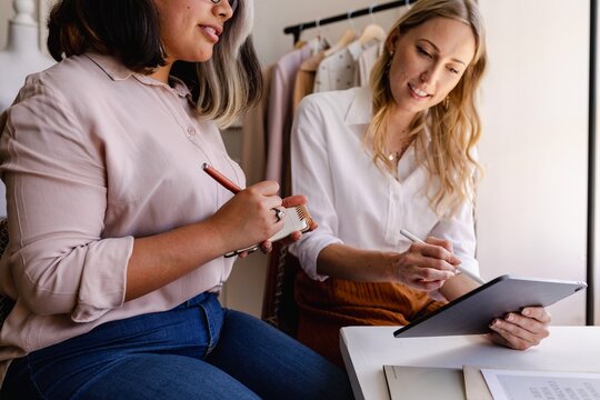 Diverse business women collaborating, writing notes, using a tablet. Business coworker women taking notes, using a tablet, collaborating. Business discussion between women. Women working together - Powered by Adobe