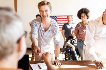 US election day, Happy man and diverse people wait in line to vote at US election station with American flag in background. Diverse people wait in line to vote at US election day. American democracy.