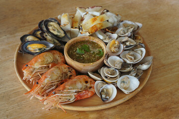 Closeup Grilled seafood, three large shrimps, cut squid, cockles, scallops and seafood dipping sauce in a wooden bowl on a wooden tray, on a wooden table background
