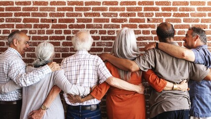 Group of diverse elderly friends, arms around each other, standing against a brick wall. Unity and friendship among seniors, showing support and togetherness. Senior diverse people holding each other.