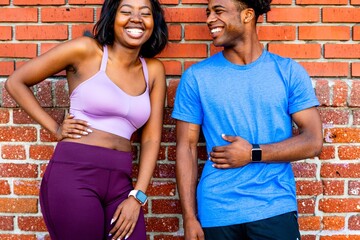 Two black people smiling, wearing casual athletic wear. Woman and man standing against a brick wall. Black couple enjoying a cheerful moment together.