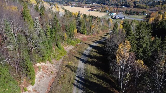 A forest path winds through a mountain ablaze with autumnal colours, leading to a few buildings. Mont-Castor, Matane, Quebec, Canada, 2025.