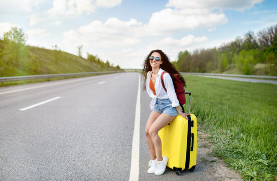 Summer adventure and autostop tourism concept. Pretty young woman in casual wear sitting on suitcase along road, waving down car, hitchhiking on roadside, having summertime trip, copy space