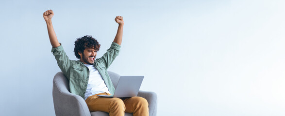 A young man sits in an armchair with a laptop on his lap. He raises his arms in joy, having achieved something significant. The setting is simple and modern, with bright light coming in.