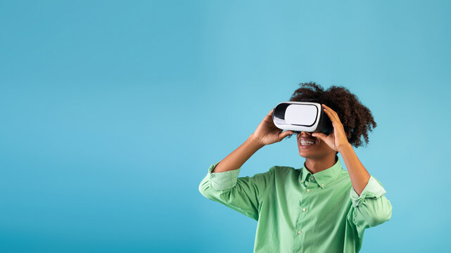 Modern gaming. African american guy wearing and touching VR headset, exploring cyberspace, using virtual reality for entertainment or studies on blue background, panorama with empty space
