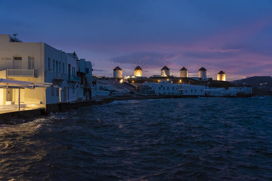 View of whitewashed buildings stand next to the dark, rippling sea under a vibrant twilight sky, with iconic windmills silhouetted against the horizon, Mykonos, Mykonos, Greece.