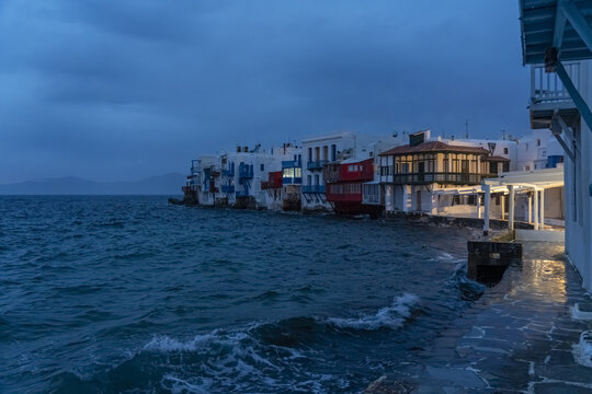 View of whitewashed buildings with colorful accents line the waterfront as waves crash against the shore in the twilight, Little venice, Mykonos, Greece.