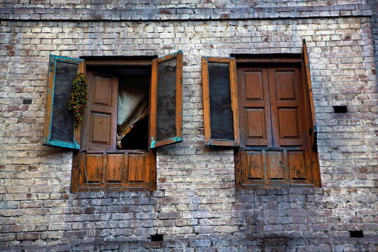 View of weathered brick wall with two rustic wooden windows, one ajar revealing a glimpse of interior, the other shut tight, Lahore, Punjab, Pakistan.