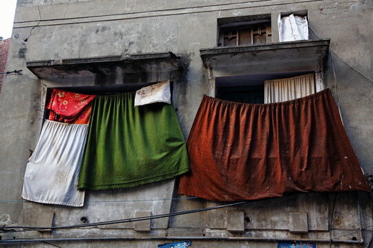 View of weathered building facade with vibrantly colored curtains hanging from windows, casting shadows on the aged walls, Lahore, Punjab, Pakistan.