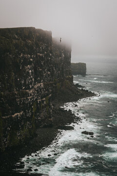 View of rugged cliffs meet the churning, foamy sea under a blanket of thick fog, a solitary figure perched at the edge, Latrabjarg, Westfjords, Iceland.