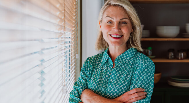 Smiling senior woman relaxing at home in kitchen by window with blinds
