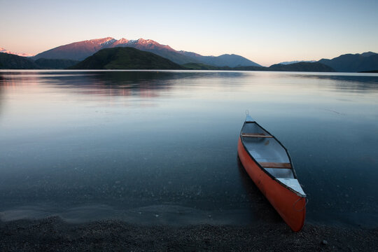 View of an orange canoe rests on the shore of a serene lake, mirroring the soft hues of the dawn sky and distant snow-capped mountains, Wanaka, Otago Region, New Zealand.