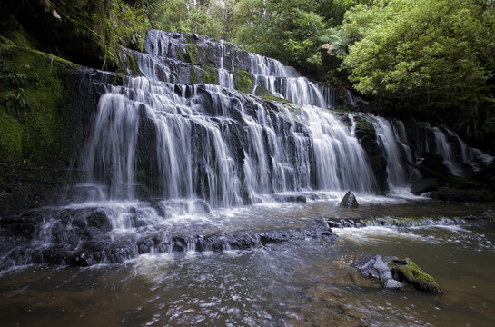 View of cascading water plunges down dark, moss-covered rocks, framed by vibrant green foliage, creating a serene scene, Purakaunui, Otago Region, New Zealand.