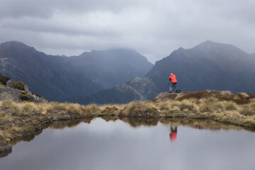 View of a lone figure in a vibrant red jacket stands poised with a camera, mirroring in the still waters against a backdrop of brooding mountains, Christchurch, West Coast Region, New Zealand.