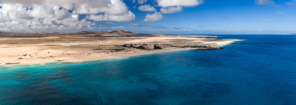 Aerial view shows Playa del Bajo de la Burra in Fuerteventura, pale gold dunes, crescent shore, turquoise shallows to deep Atlantic, volcanic hills, resorts, palms, midday light.