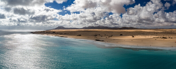 Aerial view of Fuerteventura, Canary Islands, with golden dunes, turquoise shoreline, low volcanic cones, and ridges as sunlight moves across sea and desert like plain.
