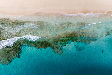 Aerial top down view shows pale sand, turquoise water, and a wide shallow reef near Playa del Bajo de la Burra or Morro Jable, with shorebreak lines and seagrass. © Aerial Film Studio