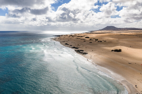 Aerial golden sand meets turquoise Atlantic waters, gentle surf, dark volcanic rocks, small beachgoers, volcanic cone near Corralejo, Playa del Bajo de la Burra.