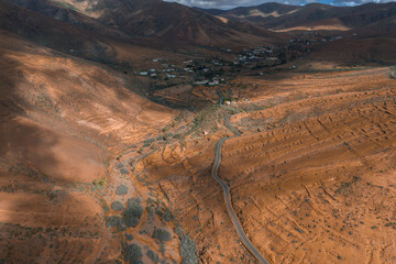 Aerial view of Fuerteventura, Canary Islands. A sinuous single lane road winds between terraced volcanic slopes, whitewashed houses cluster, clouds cast moving shadows. © Aerial Film Studio